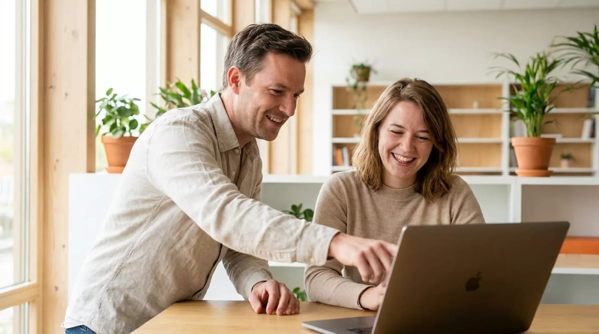 Twee collega's bekijken samen een laptop aan een houten tafel in een lichte kantoorruimte met planten.